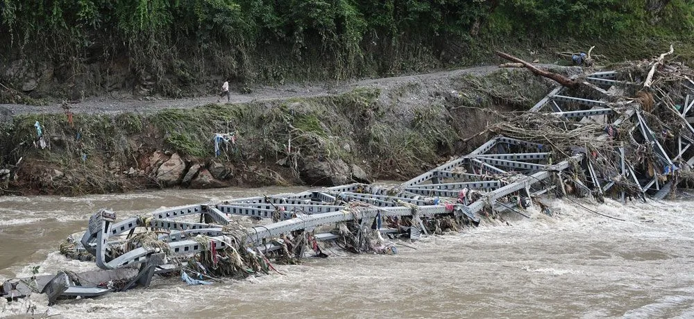 A steel truss bridge folded into a brown flooded river, with a man walking the road on the far bank