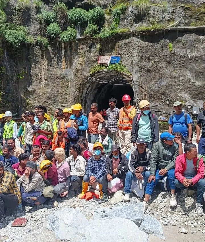 Dozens of hydropower workers in hard hats sitting and standing outside a tunnel mouth after being rescued